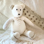 Groom bear in white wedding attire with bow tie, sitting on lace.