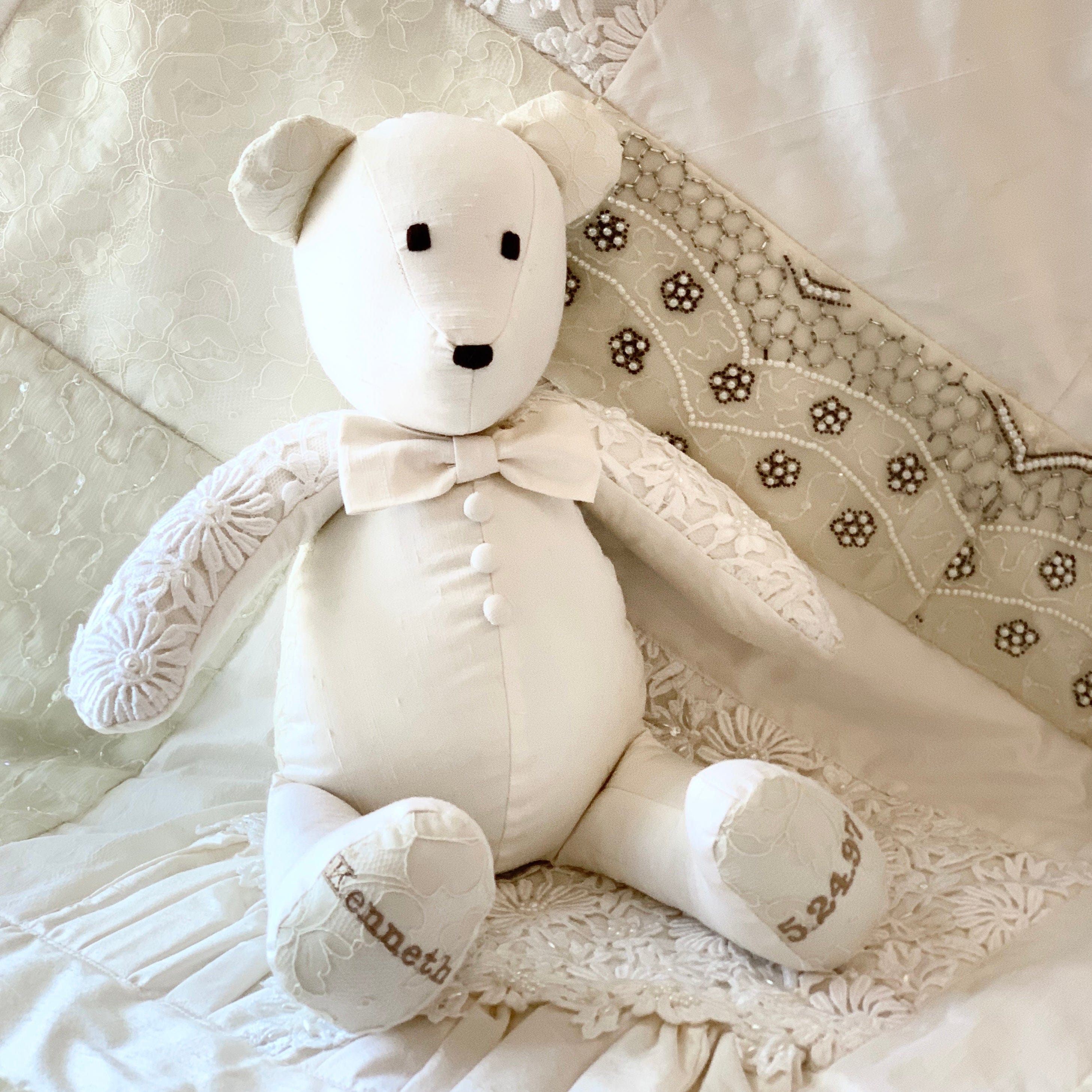 Groom bear in white wedding attire with bow tie, sitting on lace.