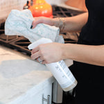 Woman cleaning kitchen counter with spray bottle and cloth.